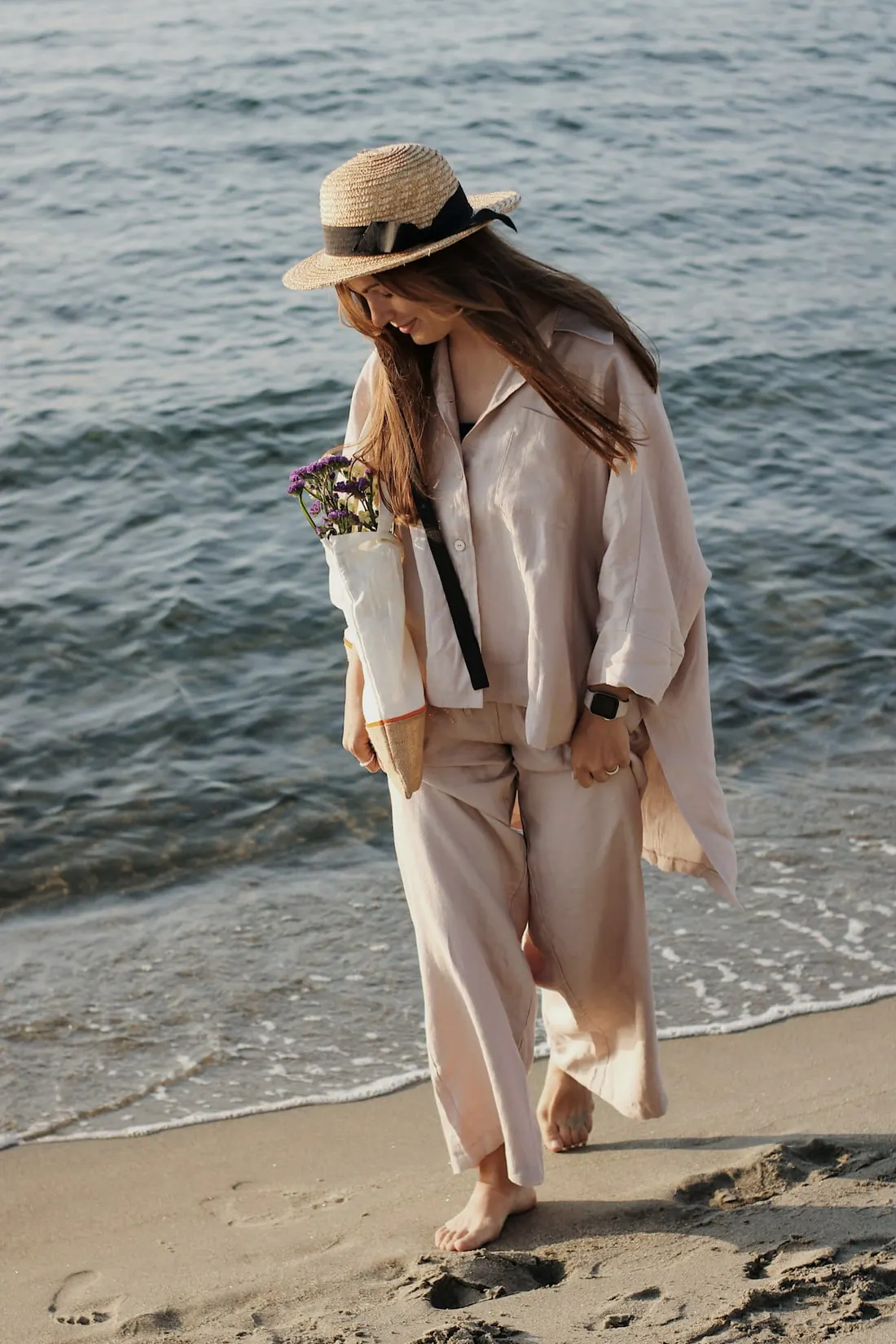 A woman in a blush linen shirt and wide trousers walking along the shoreline