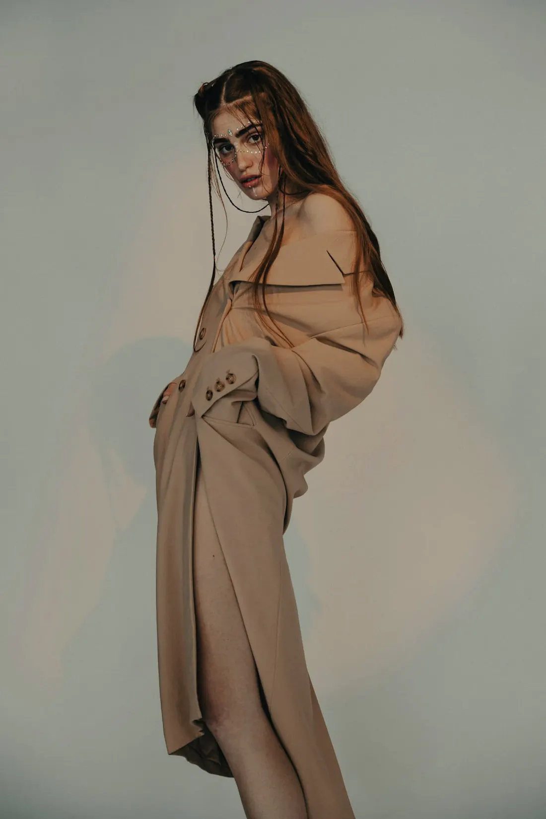A model in a camel suit against a soft sage studio wall, one hand in her hair