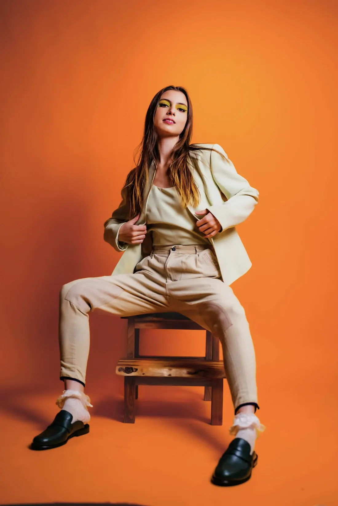 A model in a cream blazer and pleated trousers seated on a wooden stool against a saturated orange backdrop