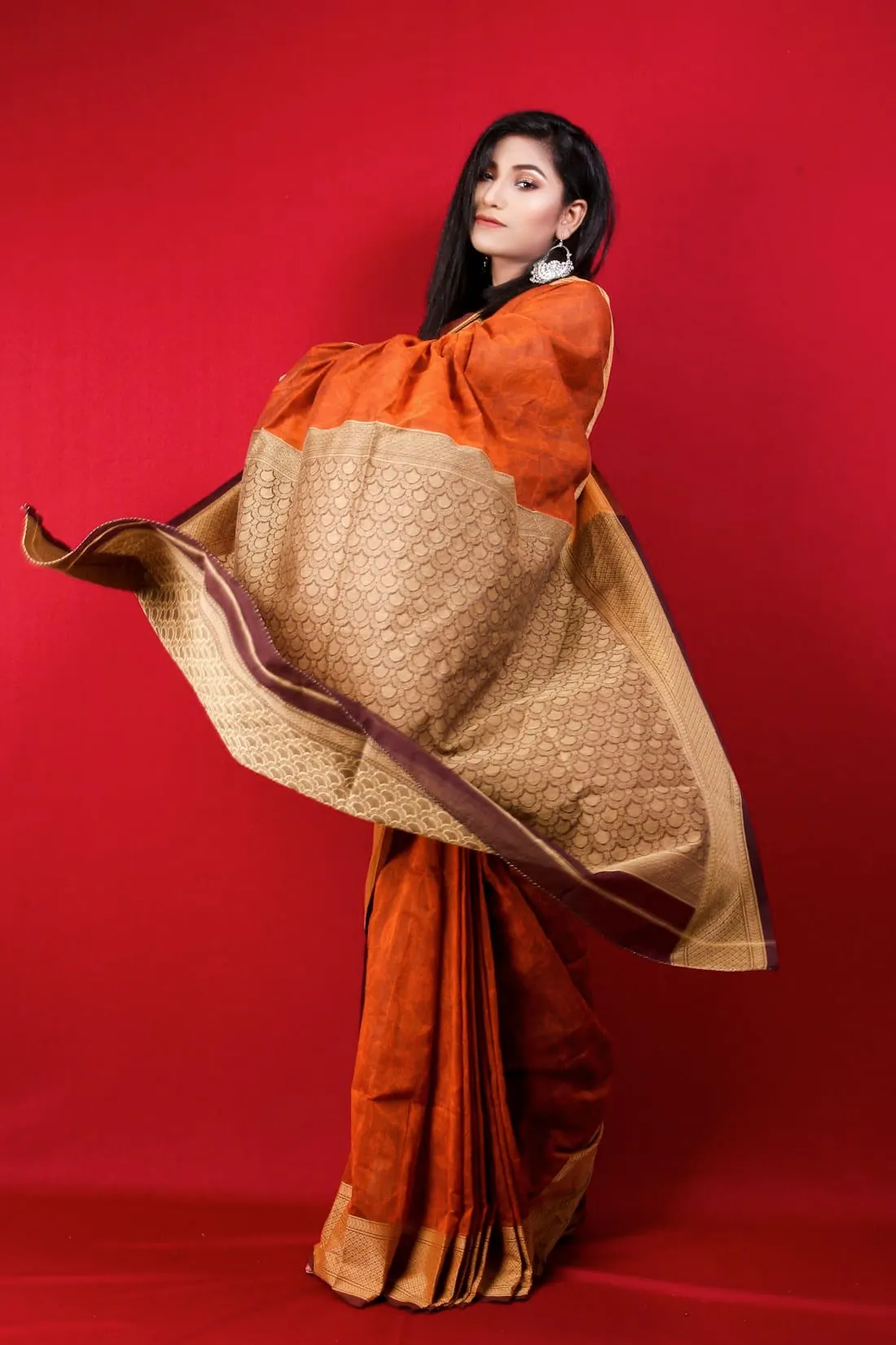A woman in a flowing burnt-orange saree against a deep red backdrop