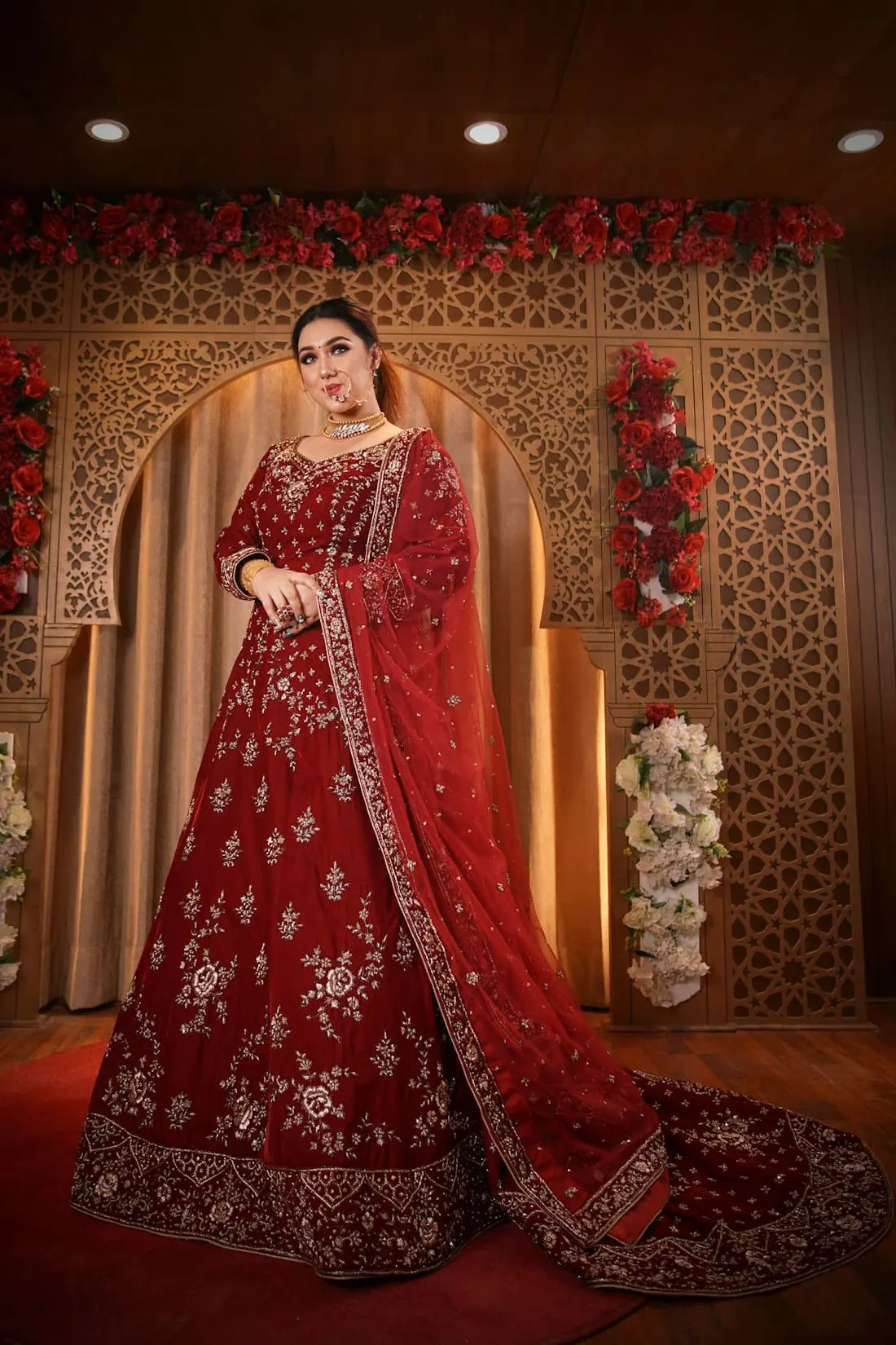 A bride in a deep red embroidered lehenga under a carved arch